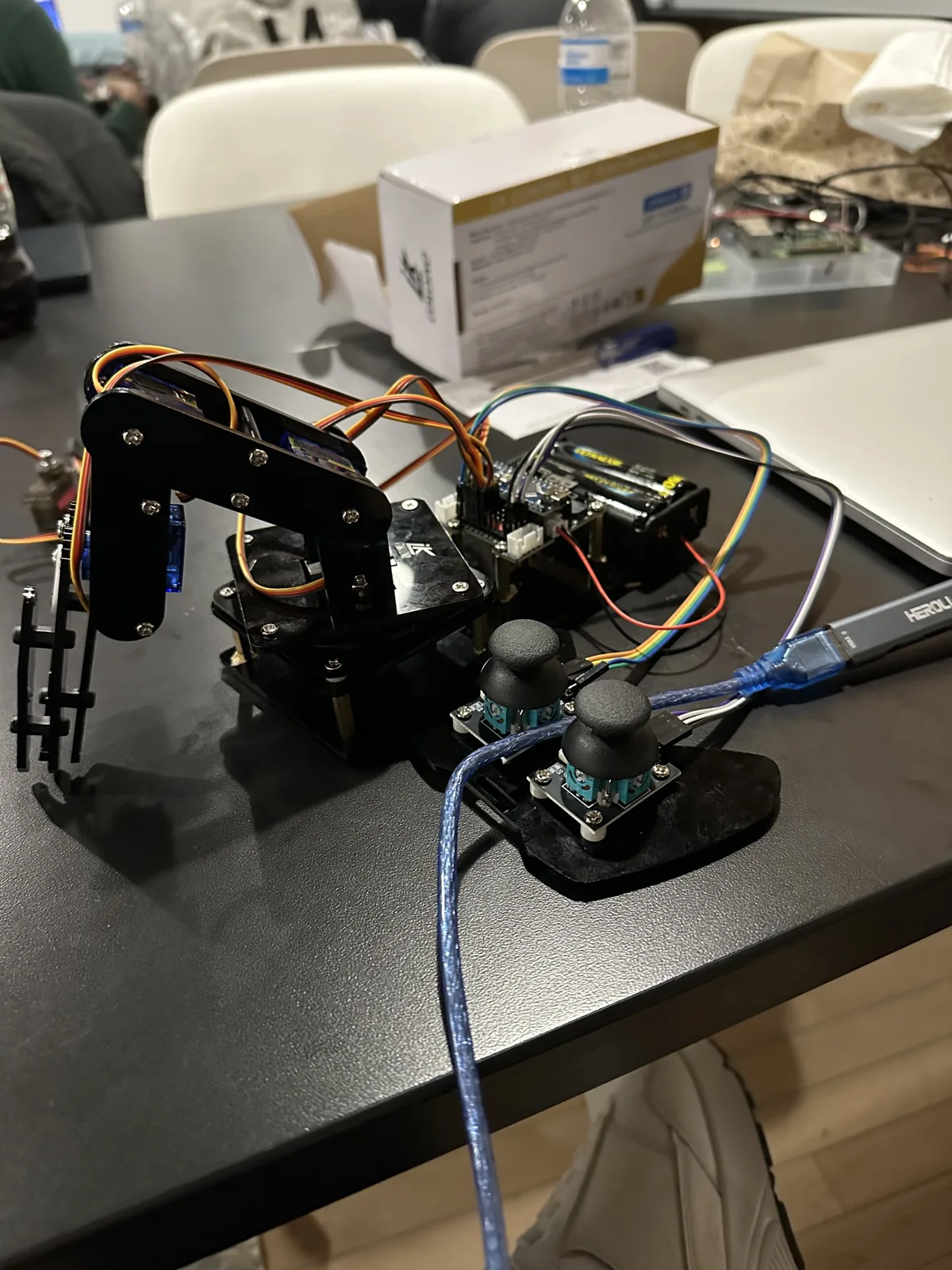 Close-up of the four-servo robot arm on a black table, with its two joysticks and wiring beside it