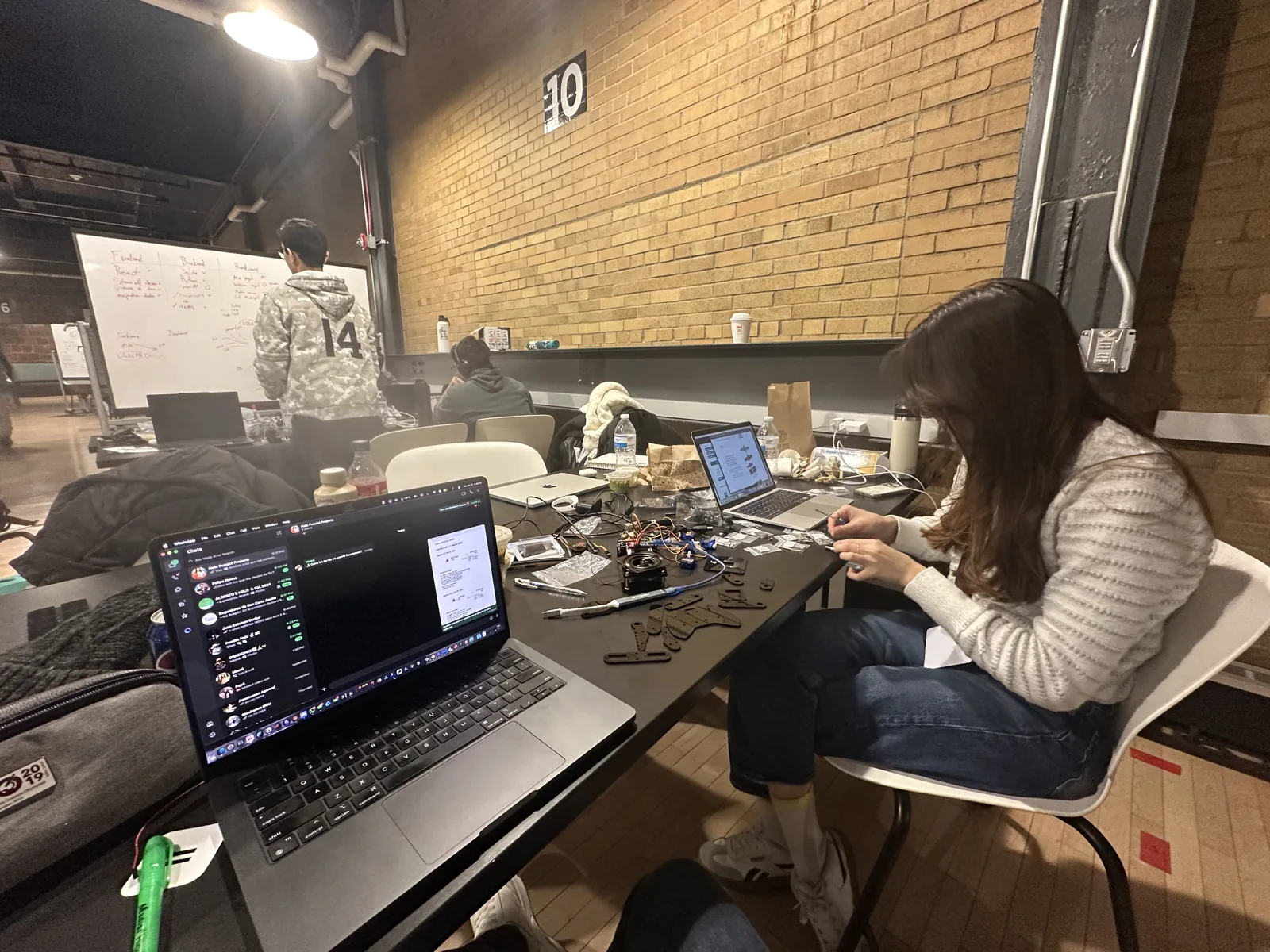 A teammate assembling the arm at a table covered in parts and laptops in a brick-walled hackathon hall at night