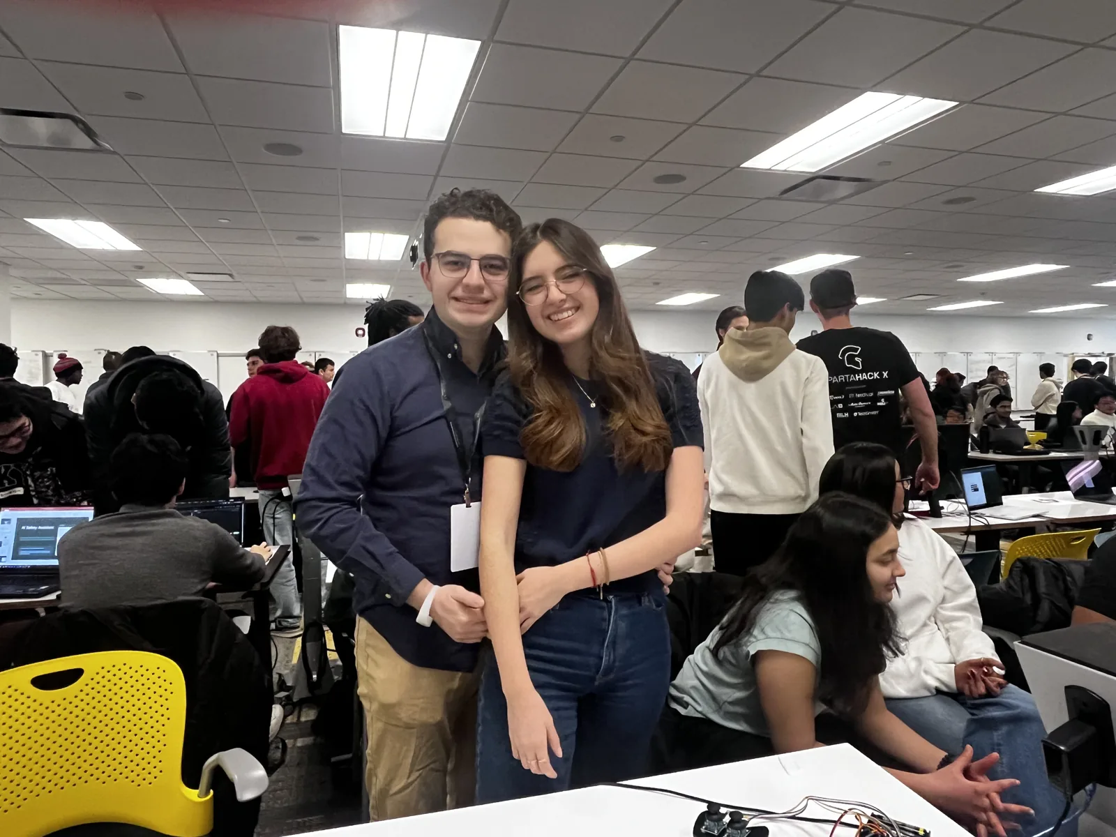 Daniel Helo Puccini and Daniela Ojeda smiling together on the SpartaHack floor, hackers at work behind them